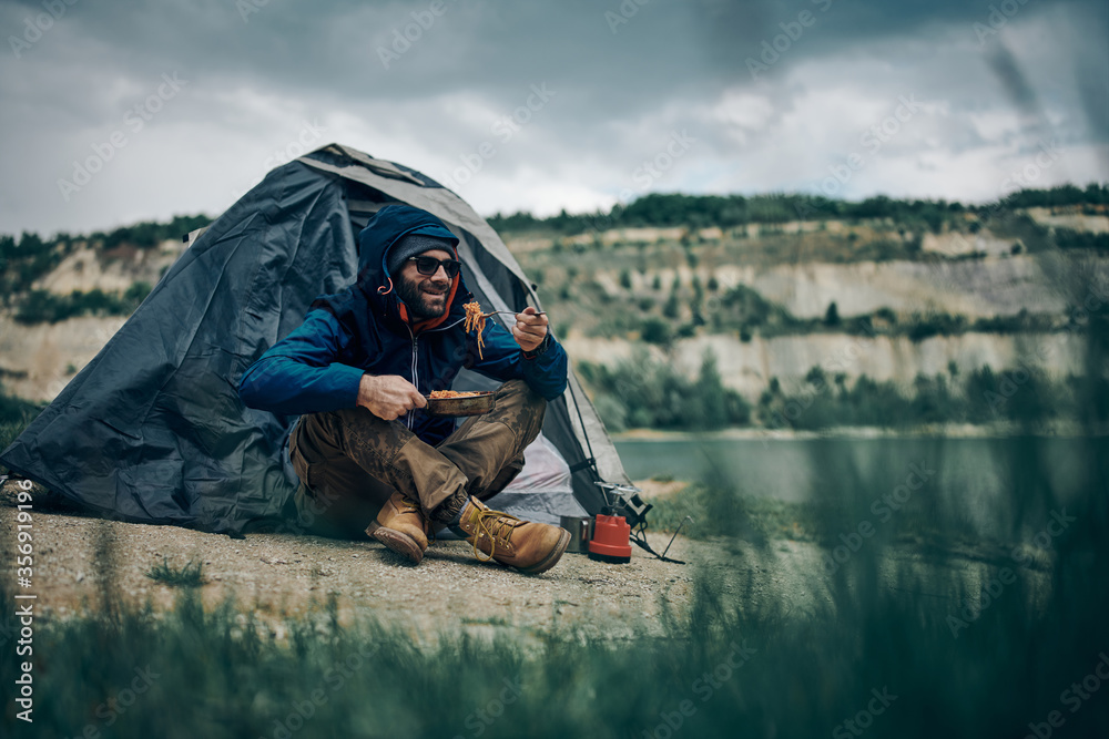 Young attractive bearded smiling man sitting by the tent on camping trip and eating his lunch. Young attractive bearded smiling man sitting by the tent on camping trip and eating his lunch.