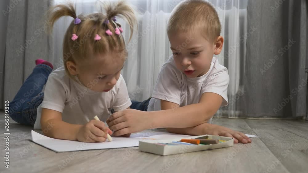 Cute children draw with crayons on white paper lying on the floor in the room.