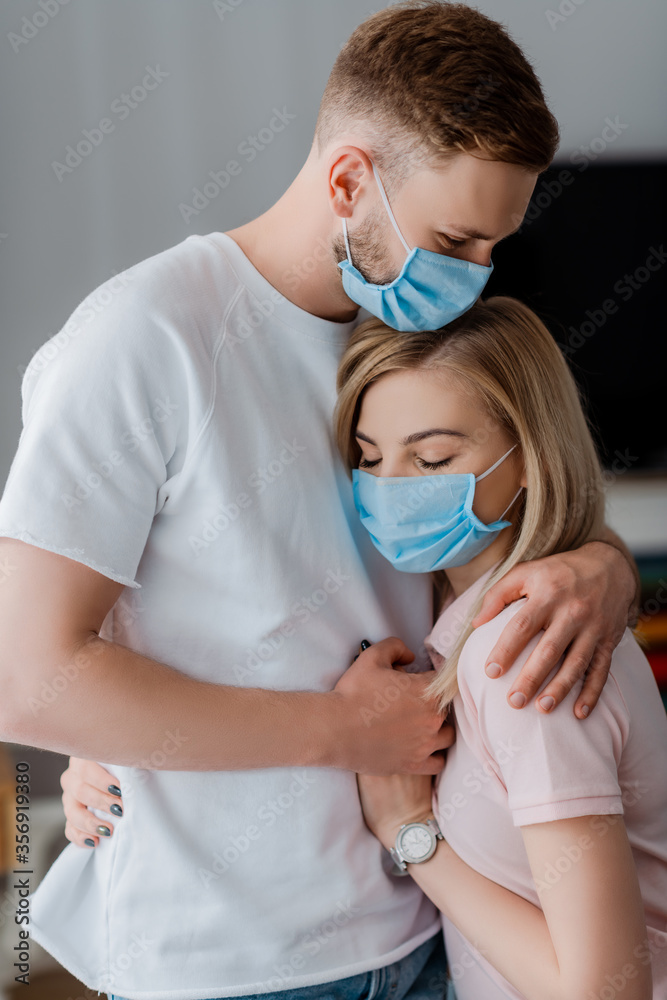 couple in medical masks holding hands and hugging at home