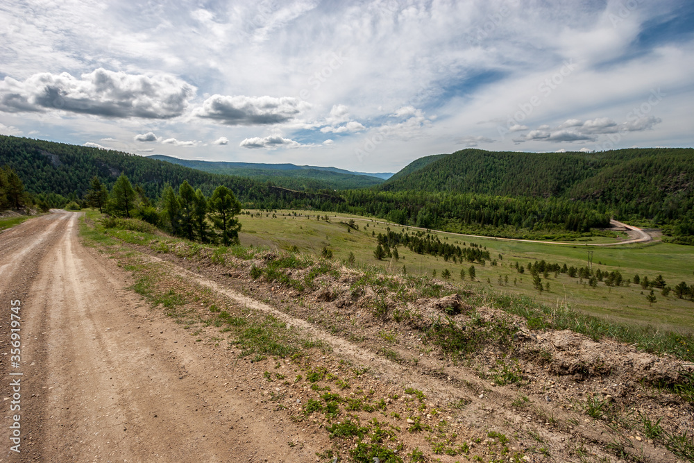 A winding road goes between green mountains with a forest. Dirt road. Clouds on the blue sky. Sunny. Horizontal.