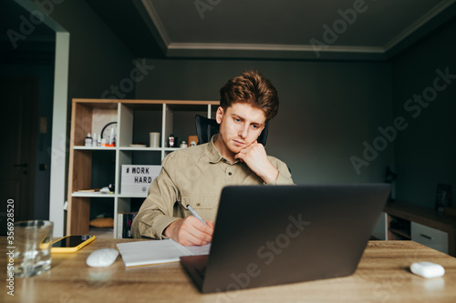 Portrait of a pensive student in a shirt sitting at home on a laptop and studying, looking at the computer and writing in a notebook. Serious student learns remotely