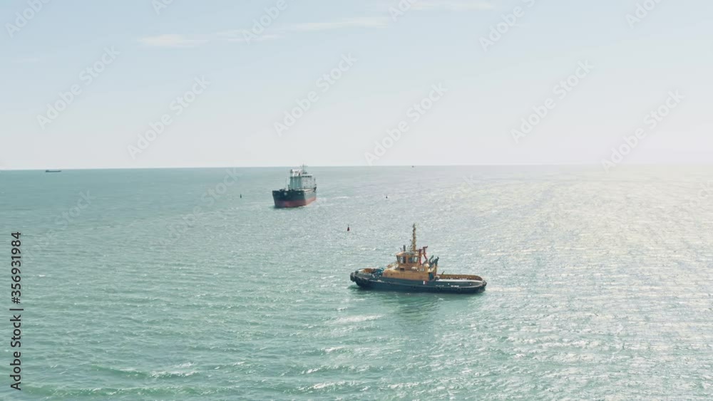 Aerial drone video of yellow tugboat goes to a large cargo ship to transport to the dock. Cargo ship barge and tugboat sail to meet each other in the seaport of the port. Aerial View. Drone shoot. Aerial drone video of yellow tugboat goes to a large cargo ship to transport to the dock. Cargo ship barge and tugboat sail to meet each other in the seaport of the port. Aerial View. Drone shoot.