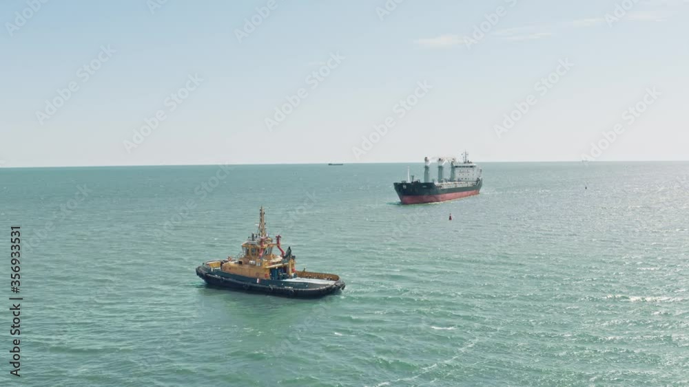 Aerial drone video of yellow tugboat goes to a large cargo ship to transport to the dock. Cargo ship barge and tugboat sail to meet each other in the seaport of the port. Aerial View. Drone shoot. Aerial drone video of yellow tugboat goes to a large cargo ship to transport to the dock. Cargo ship barge and tugboat sail to meet each other in the seaport of the port. Aerial View. Drone shoot.
