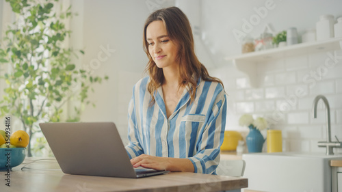 Young Beautiful Woman Using Laptop Computer While Wearing Blue Pyjamas. Brunette Female Sitting in a Modern Kitchen Room. Working from Home During Quarantine.