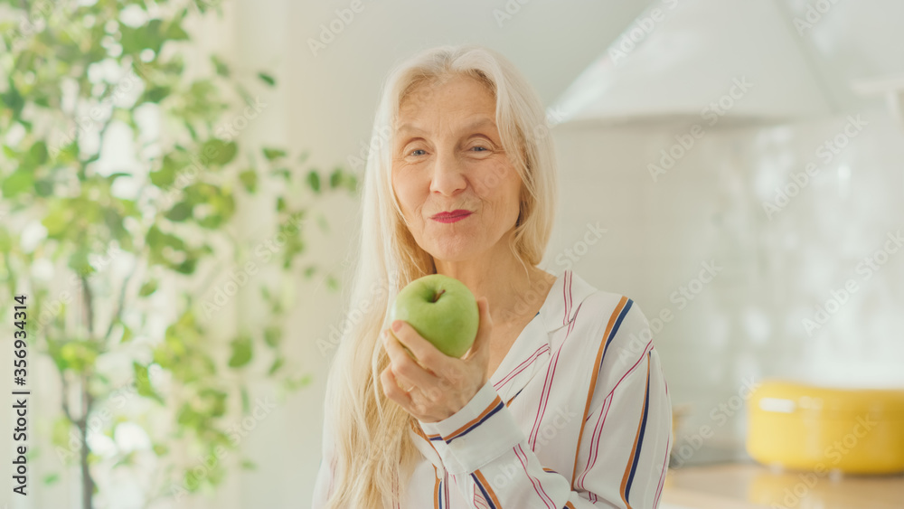 Authentic Portrait of Senior Female Pensioner in a Bright Kitchen at Home Eating an Apple. Beautiful Old Female with Gray Hair Poses for the Camera and Gently Smiles. Happy Vegetarian Full of Health. Authentic Portrait of Senior Female Pensioner in a Bright Kitchen at Home Eating an Apple. Beautiful Old Female with Gray Hair Poses for the Camera and Gently Smiles. Happy Vegetarian Full of Health.