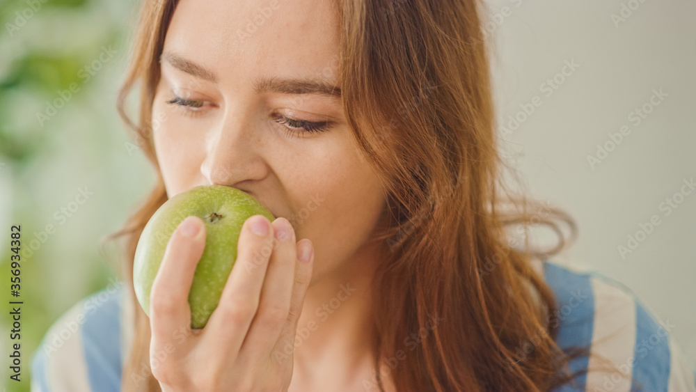Authentic Portrait of a Beautiful Young Female in a Bright Room at Home Eating an Apple. Attractive Brunette Woman Poses for the Camera and Gently Smiles. Happy Vegetarian Full of Health. Authentic Portrait of a Beautiful Young Female in a Bright Room at Home Eating an Apple. Attractive Brunette Woman Poses for the Camera and Gently Smiles. Happy Vegetarian Full of Health.