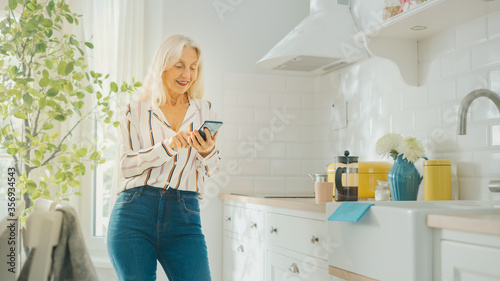 Authentic Senior Female in Blue Jeans and White Striped Blouse is Using Smartphone in the Kitchen. Happy Beautiful Pensioner Relaxing at Home.