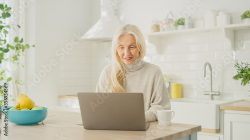 Authentic Senior Woman Using Laptop Computer in a Bright Kitchen Room at Home. Beautiful Old Female Pensioner with Gray Hair Browses Internet on a Computer. Happy Elderly Person Full of Health.