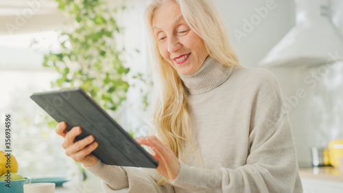 Authentic Senior Woman Using Tablet Computer in Kitchen Room at Home. Beautiful Old Female Pensioner with Gray Hair Writes a Text in a Messenger on a Device. Elderly Person in Hand with Technology.