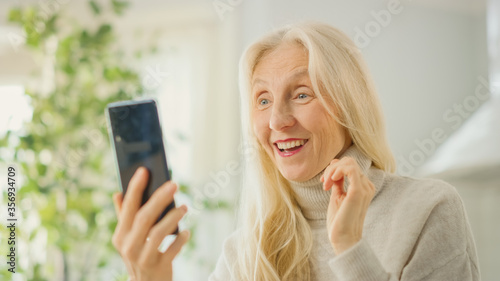 Authentic Senior Woman Answering a Video Call Using Her Smartphone in Kitchen Room at Home. Beautiful Old Female Pensioner with Gray Hair is Happy to See Her Family on Video Chat.