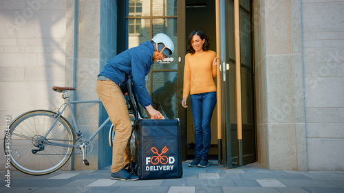 Happy Food Delivery Man Wearing Thermal Backpack on a Bike Delivers Restaurant Order to a Happy Female Customer. Courier Delivers Takeaway Lunch to Office Building. © Gorodenkoff