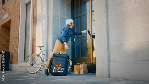 Food Delivery Man Wearing Thermal Backpack on Bike Delivers Restaurant Order, Leaving it Under the Door. Concept: Contactless (No Contact) Delivery for Social Distancing, Self Isolation
