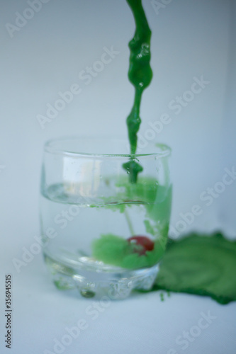 In a transparent glass with clean water, with cherries at the bottom, green paint is poured with a dynamic movement and colors the water green on a white background.