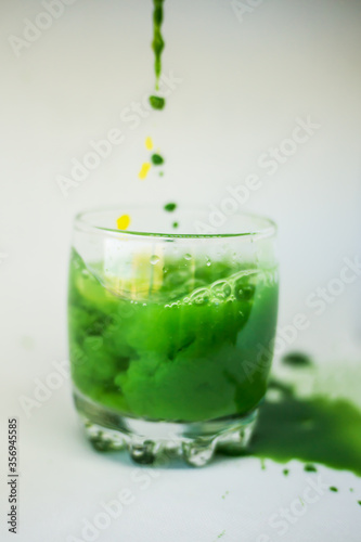 Green and yellow paint is poured into a transparent glass with pure water in motion and paints transparent water in green and yellow on a white background.