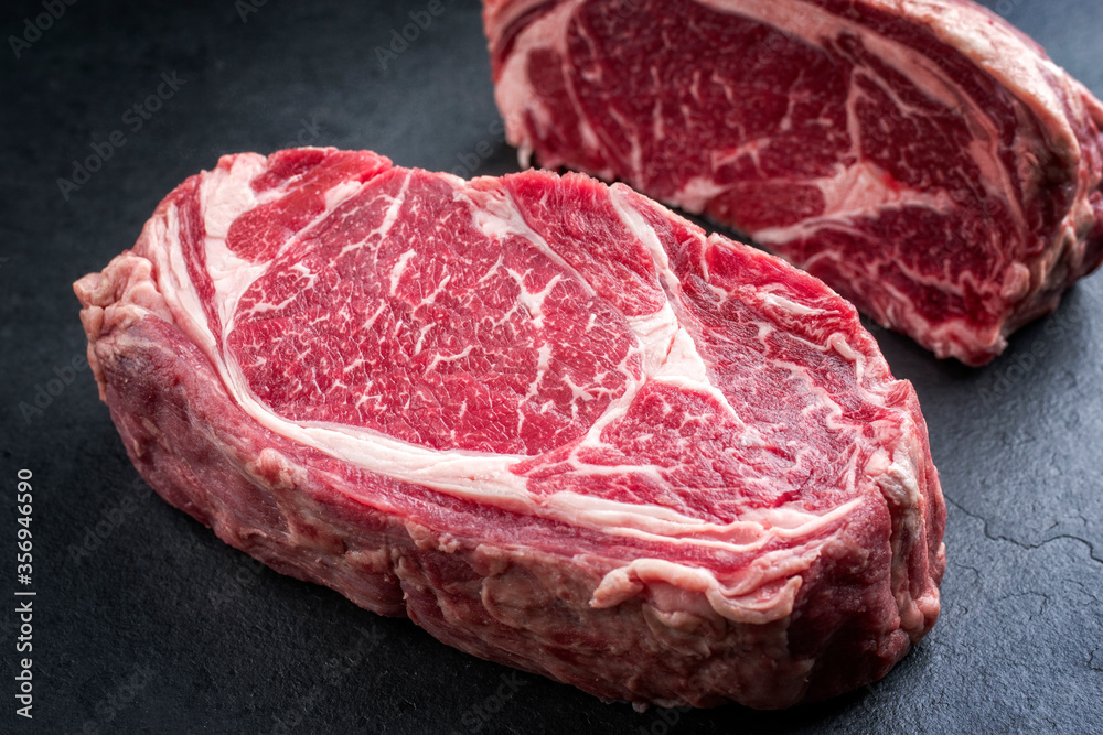 Obraz premium Two raw dry dged Kobe rib-eye steaks as closeup on a black board