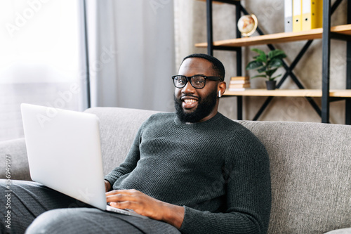 Cheerful African-American guy sits on sofa in modern living room alone and using trendy laptop for net searching, web surfing or texting messages. A black guy in glasses looks into a camera and smiles