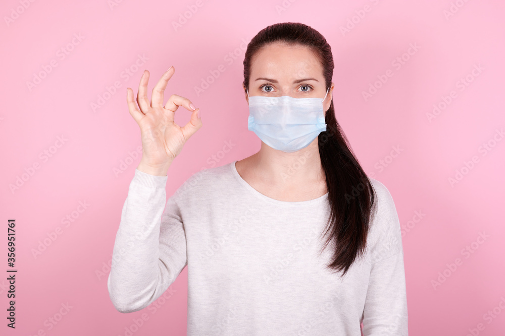 Portrait of a brunette girl in a medical mask shows OK sign with hand.