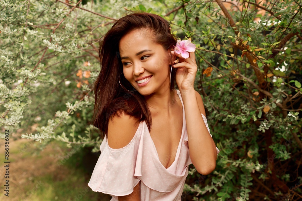 Summer portrait of lovely Asian woman with flower in hairs posing in the garden. Natural make up, round earrings and pink dress. Summer portrait of lovely Asian woman with flower in hairs posing in the garden. Natural make up, round earrings and pink dress.