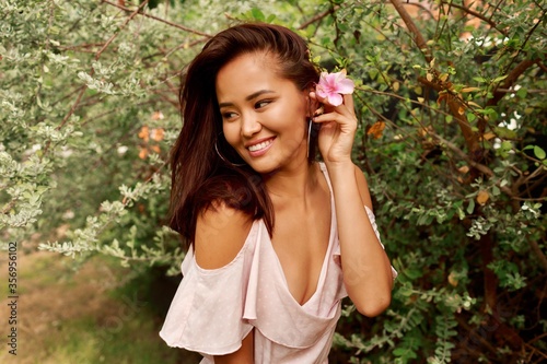 Summer portrait of lovely Asian woman with flower in hairs posing in the garden. Natural make up, round earrings and pink dress.