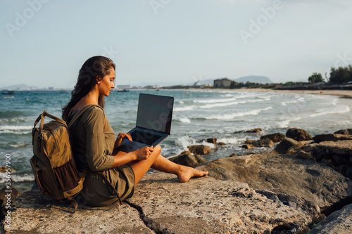 Young pretty female freelancer working on a laptop while traveling by the sea, remote work concept. Work by the sea