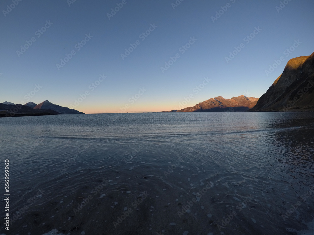 beautiful calm blue waves hitting white frozen sandy beach in late autumn in the arctic circle with deep mountain and open sea view on clear sky background