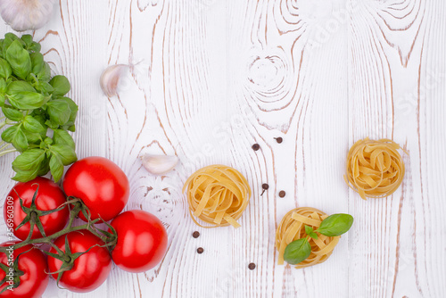 Top view raw tagliatelle pasta with fresh basil, garlic and tomatoes on a rustic white table, flat lay, copy space.