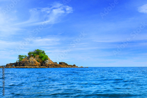 Beautiful view of blue sea around the rock island with white cloud in bright blue sky. Bushes and green tree grow at the top of island around by blue sea in sunny day of summer