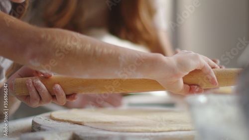 Woman teaching girl to roll dough on kitchen table. Girl hands using rolling pin