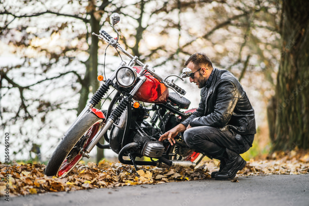 Bearded brutal man in sunglasses and leather jacket sitting on a motorcycle on the road in the forest