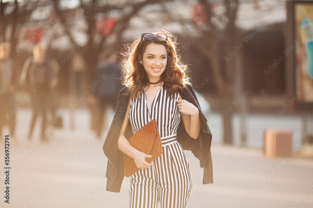 Fototapeta premium Beautiful brunette woman in stylish spring look, wearing in striped suit and black jacket, holding red handbag, Smiling girl with make up after beauty salon walking at street, posing at camera.Sunset.