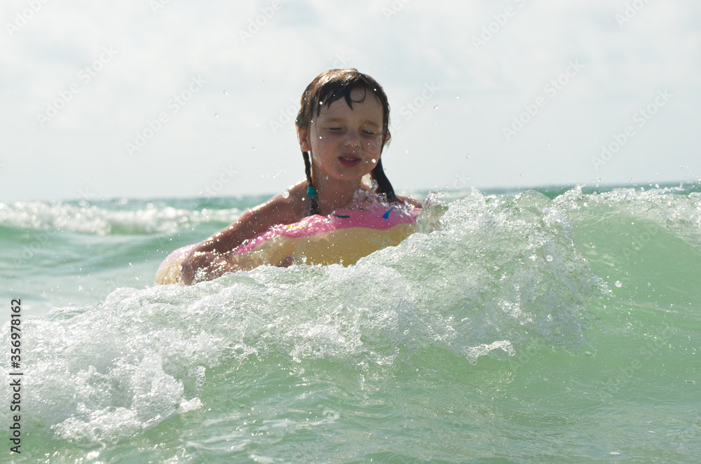 A little girl in a swimming circle is swimming in the sea. The child has fun on vacation. Sea foam and wave splashes