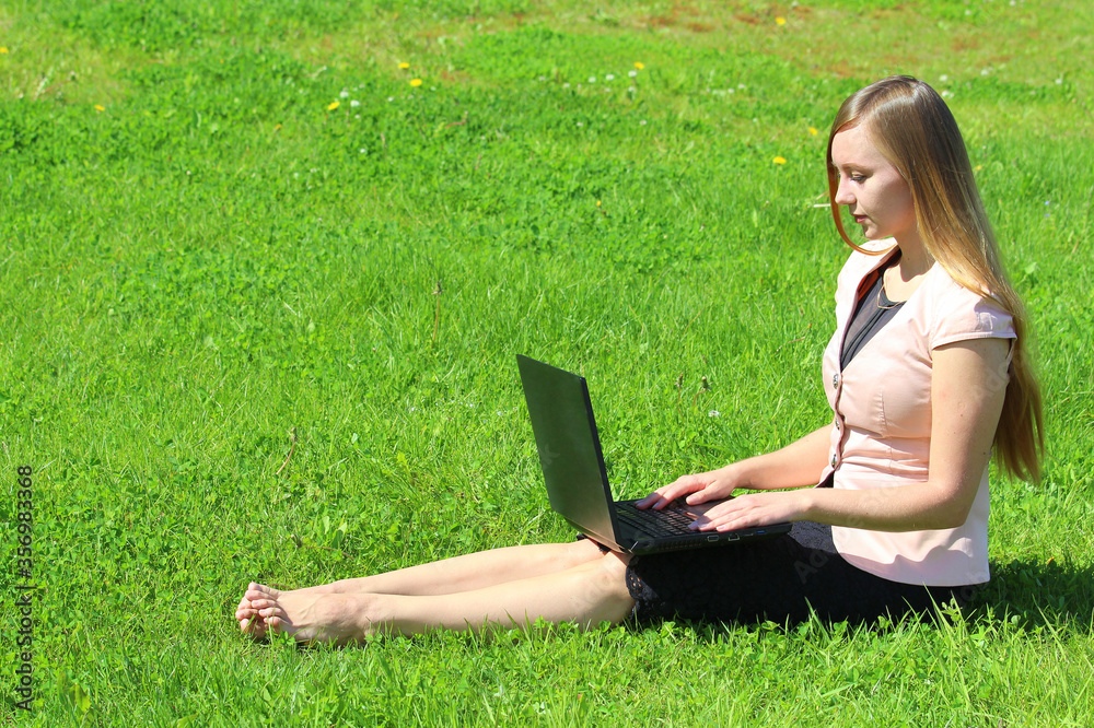 A beautiful young white girl in a pink jacket and black skirt and with long hair sitting on green grass, on the lawn and working behind a black laptop A beautiful young white girl in a pink jacket and black skirt and with long hair sitting on green grass, on the lawn and working behind a black laptop