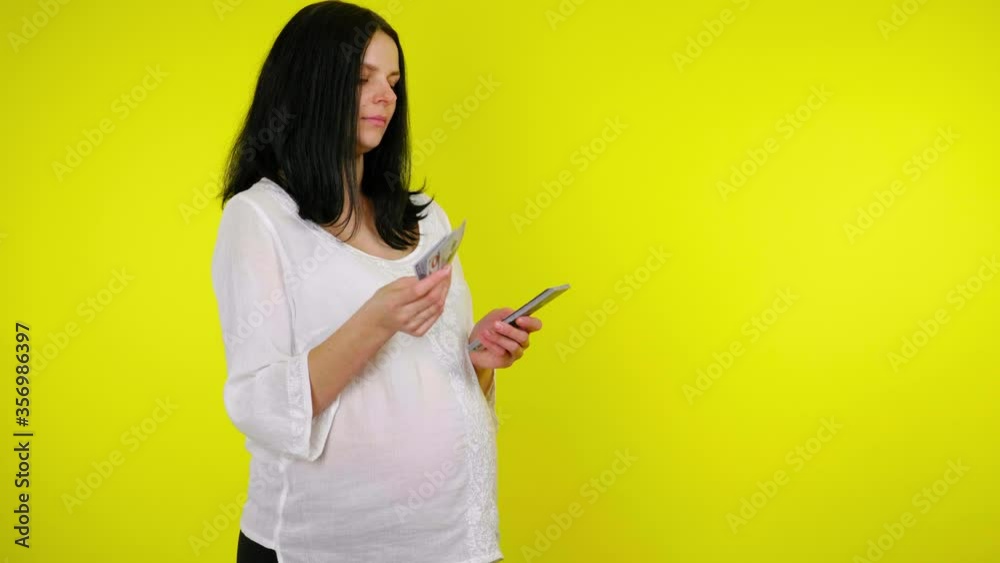 Rich young pregnant woman with black hair in a white blouse counts money on a yellow background with copy space. Place for text or product. 4K slow motion footage Rich young pregnant woman with black hair in a white blouse counts money on a yellow background with copy space. Place for text or product. 4K slow motion footage