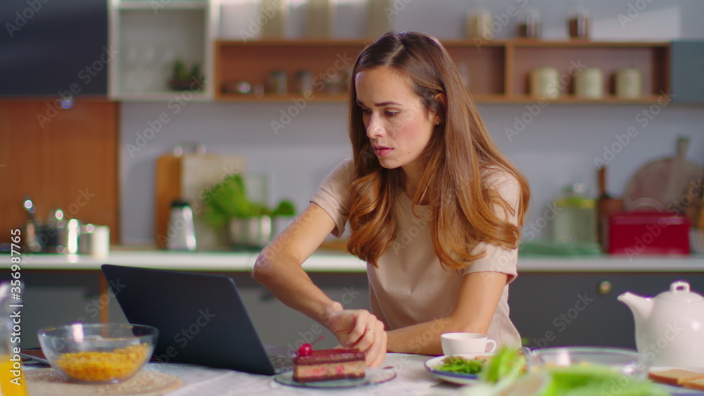 Businesswoman working on laptop at home office. Girl eating cake on kitchen