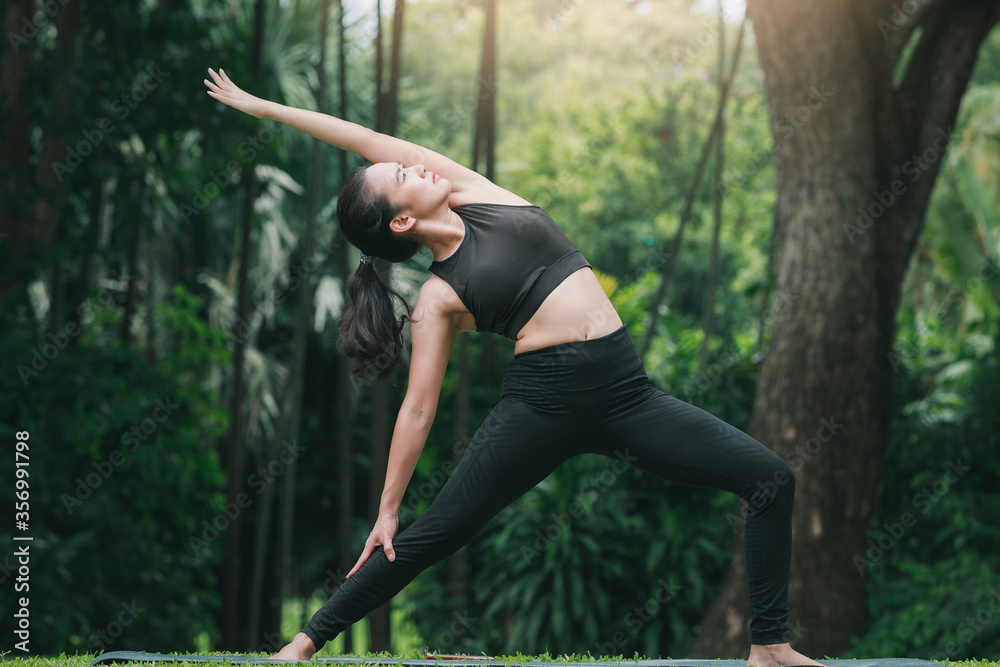 Fototapeta premium Asian woman practicing yoga in Warrior Pose (Virabhadrasana) on the mat in outdoor park.