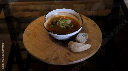 Bouillabaisse soup rotates on the wooden plate on dark tabe. Menu presentation for the restaurant. Seafood soup contains hot spicy broth, squid, shrimps, shells, parsley. 