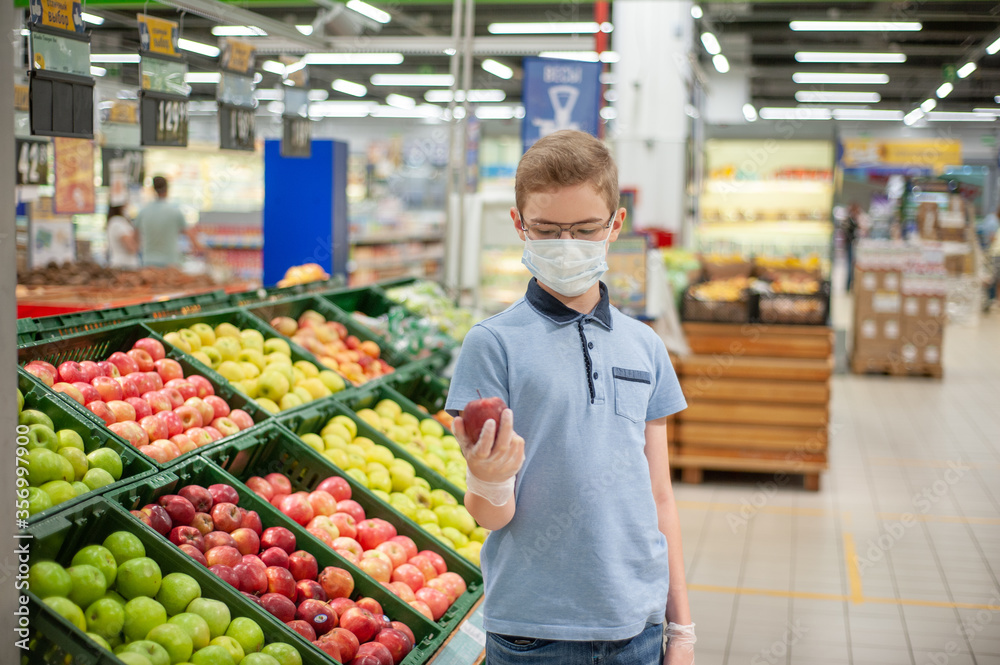 A teenager in a supermarket holding an apple. He's wearing a mask and gloves.