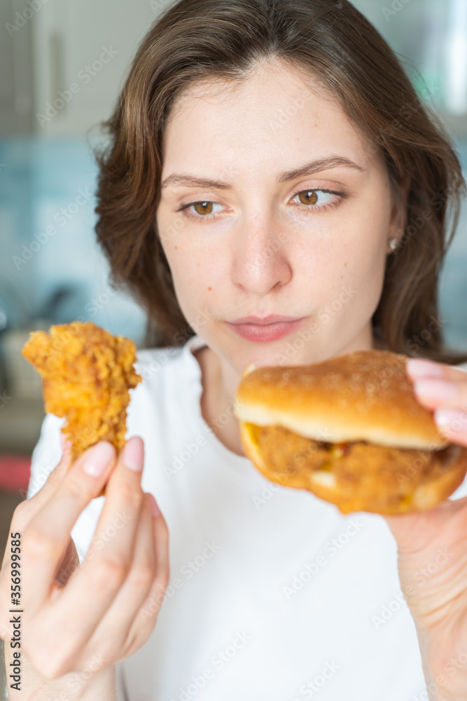 Hungry woman chooses what to eat first. Harmful cheeseburger and chicken wing in female hands Hungry woman chooses what to eat first. Harmful cheeseburger and chicken wing in female hands