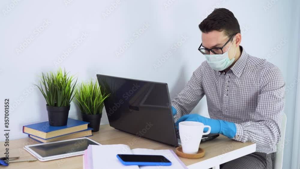 Male brunette businessman wear plaid shirt, medical disposable face mask, blue gloves, black glasses. Young man works at computer, answers phone call, drink coffee. On table books, notebook, tablet. Male brunette businessman wear plaid shirt, medical disposable face mask, blue gloves, black glasses. Young man works at computer, answers phone call, drink coffee. On table books, notebook, tablet.