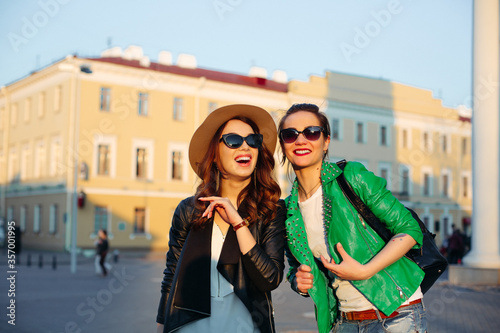 Two beautiful and shocked brunette girls stylish wearing walking at street, pointing by finger away and surprised looking with opened mouth. Emotionally girlfriends in sunglasses, hat, leather jacket. © Вячеслав Косько