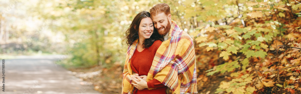 Fototapeta premium Beautiful couple man woman in love. Boyfriend and girlfriend wrapped in yellow blanket hugging in park on autumn fall day. Authentic real people. Banner header for a website.