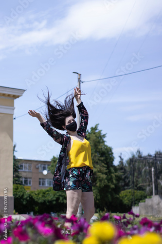Attractive young brunette woman in yellow t-shirt and flowery shorts costume. In medical black face mask. modern reality. near bushes with flowers. covid-19 concept. copy space. High quality photo