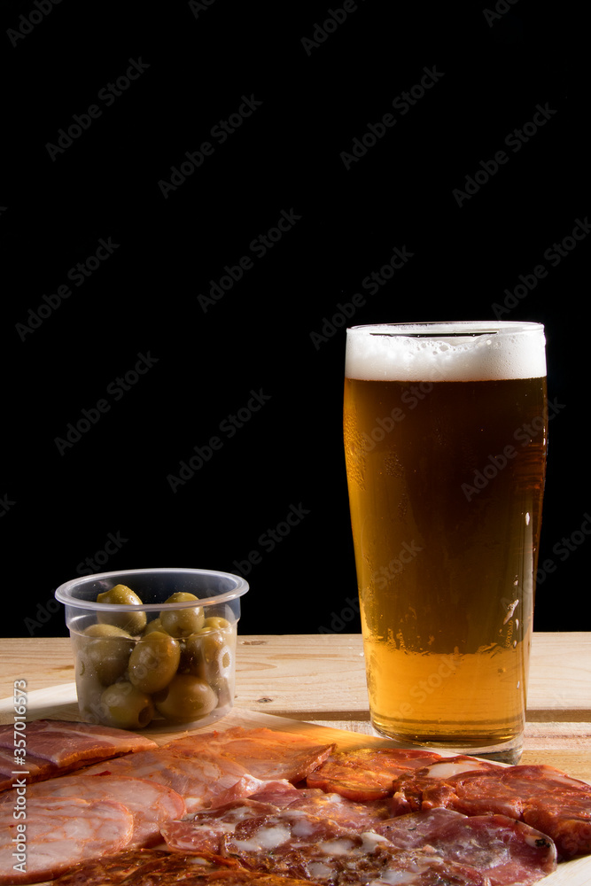 A cold glass of craft beer with a portion of snacks of cold cuts with olives, parma ham and salamis on wooden table over black background. Happy hour time. A cold glass of craft beer with a portion of snacks of cold cuts with olives, parma ham and salamis on wooden table over black background. Happy hour time.