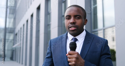 Portrait of young African American handsome male journalist talking with microphone for news episode outdoor. Pandemic concept. Close up of man correspondent in suit and tie and with mic.