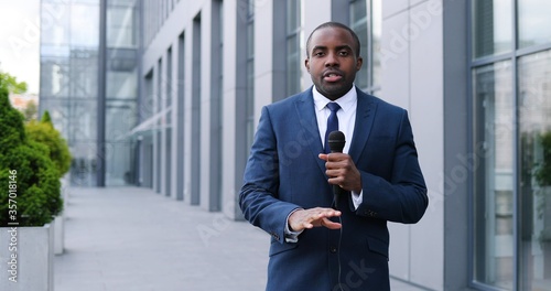 Portrait shot of young African American handsome male journalist talking with microphone for news episode outdoor. Pandemic concept. Man correspondent in suit and tie and with mic.