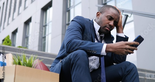 African American businessman sitting on stairs outdoors with box of personal office stuff. Angry despaired male lost work place and texting message on smartphone. Fired man scrolling, tapping on phone photo