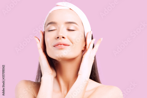Young beautiful woman with closed eyes does self-massage of face on pink background.