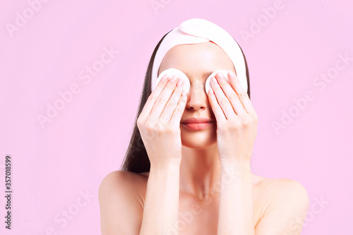 Young beautiful woman with clean perfect skin removes eye makeup with cotton pads on pink background