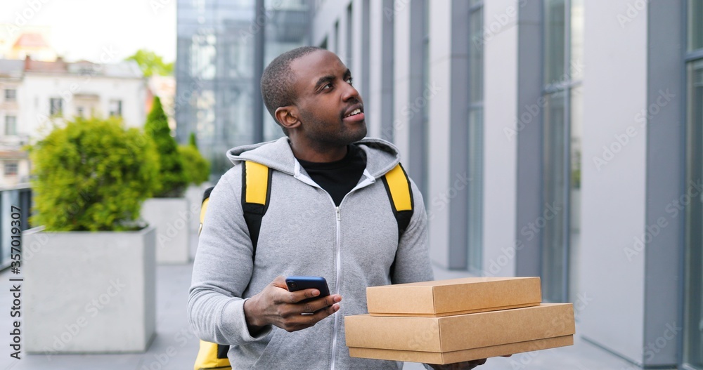 Fototapeta premium African American deliveryman with carton parcels standing the street, tapping on navigation map on phone looking for address. Male courier using navigating system on smartphone to deliver boxes.