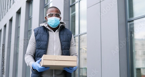 Portrait shot of handsome African American deliveryman in goggles, gloves and medical mask standing at street and holding carton boxes, giving to camera. Male courier handing pizza to client.